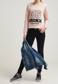 Felpa rosa con testo nero "56 BRONX MANHATTAN," jeans neri e sneakers con motivo. Tiene una giacca di jeans blu.