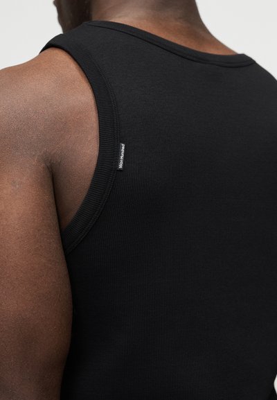 Κοντινό πλάνο στο πάνω μέρος της πλάτης και τον ώμο ενός ατόμου που φοράει μαύρο ribbed αμάνικο τοπ με μικρή ετικέτα "Won Hundred" κοντά στην περιοχή της μασχάλης.