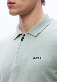 Κοντινό πλάνο ενός ατόμου που φοράει ανοιχτό πράσινο πόλο με φερμουάρ, γιακά και λογότυπο "BOSS" στο αριστερό στήθος.