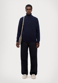 Maglione a collo alto colore navy in tessuto lavorato a maglia, abbinato a pantaloni larghi scuri e sneakers bianche, con una borsa a spalla grigia.
