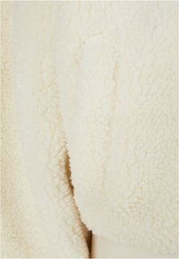Tessuto in pile di colore crema con una superficie texturizzata e soffice. Presenta un dettaglio con tasca laterale, evidenziando un aspetto morbido e accogliente.
