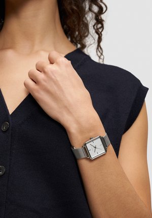 Zilveren vierkante horloge met een mesh-band, voorzien van een witte wijzerplaat en zwarte uurmarkeringen, gedragen aan een hand tegen een donkerblauwe gebreide top.