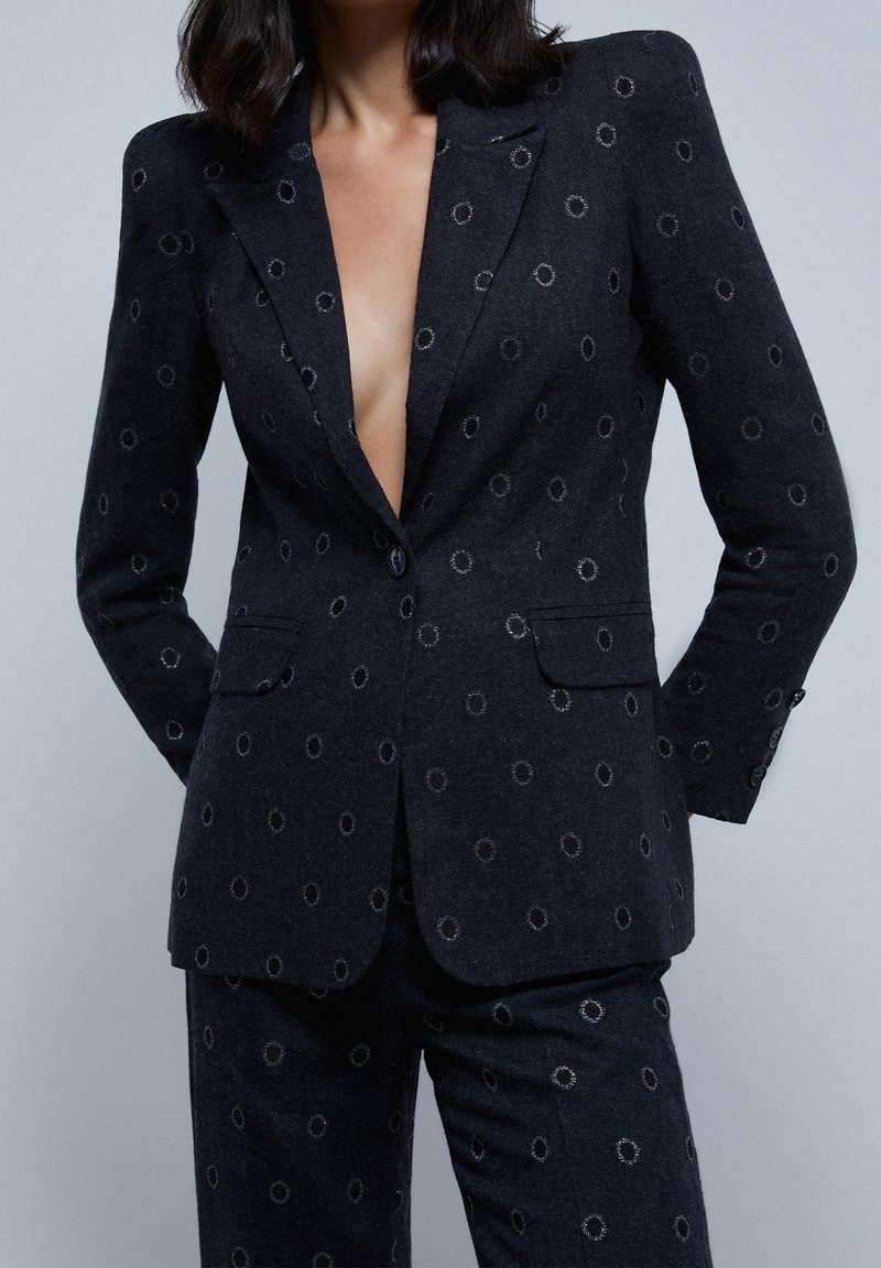 Femme portant un costume charbon foncé avec des motifs circulaires, blazer boutonné une fois, pas de chemise visible en dessous, debout les mains dans les poches.