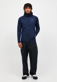 Tumed zip-up jakk, millel on vormitud disain, kõrge krae ja logo aksent. Kombineeritud mustade cargosärkide ja spordijalatsitega.