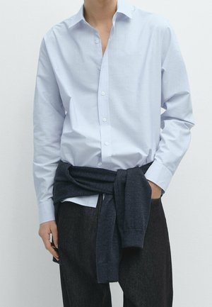Homme portant une chemise bleu clair boutonnée, un pull foncé noué autour de la taille et un pantalon foncé, debout devant un fond uni.