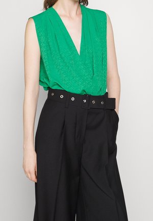 Femme portant un haut vert vif sans manches à col en V, rentré dans un pantalon noir taille haute à jambes larges, avec une large ceinture noire à œillets argentés.