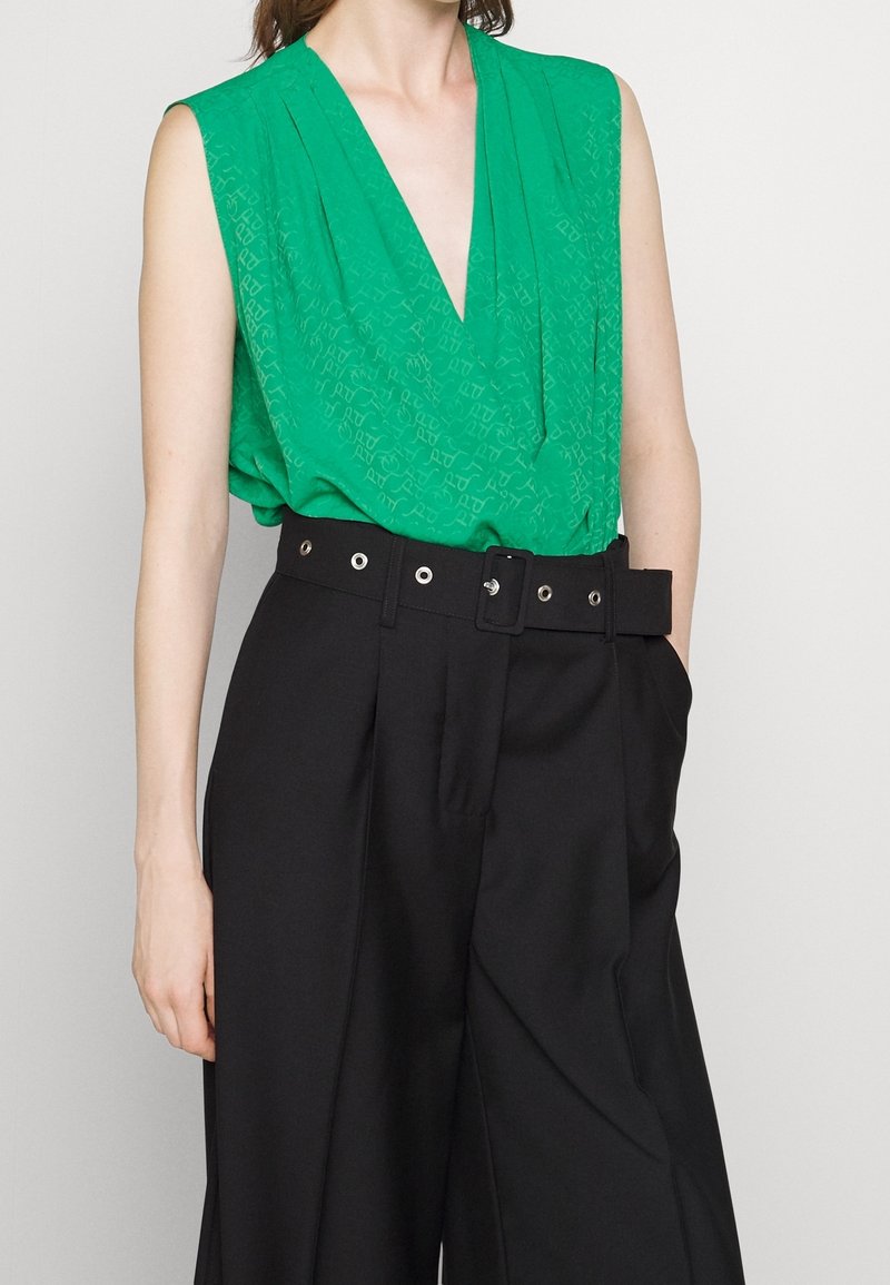 Femme portant un haut vert vif sans manches à col en V, rentré dans un pantalon noir taille haute à jambes larges, avec une large ceinture noire à œillets argentés.