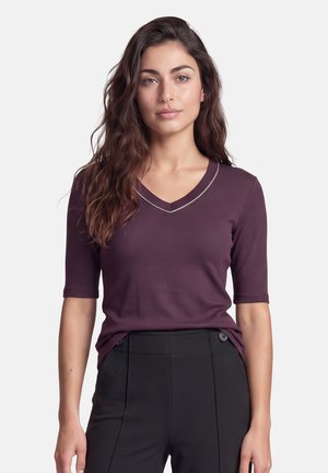 Frau mit langen, welligen Haaren, die ein bordeauxrotes V-Ausschnitt-Oberteil und hoch taillierte schwarze Hosen trägt und vor einem schlichten Hintergrund nach vorne blickt.