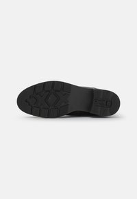 Suela de zapato negra con profundos surcos y patrones geométricos, fotografiada contra un fondo blanco liso.