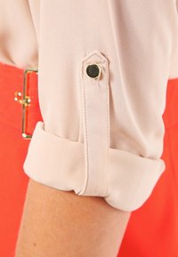 Blusa di un delicato rosa chiaro con maniche arrotolate fissate da bottoni neri, abbinata a uno sfondo rosso brillante e dettagli in hardware dorato.