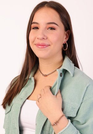 Zilveren choker met twee in elkaar verweven ringen, bijpassende armband, gedragen met een lichtgroene corduroy jasje. Oorbellen met subtiele accenten.