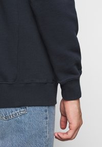 Marinblå sweatshirt med ribbade ärmar och nederkant, med en slät textur. Bärs med ljusblå denimjeans som visar en avslappnad passform.
