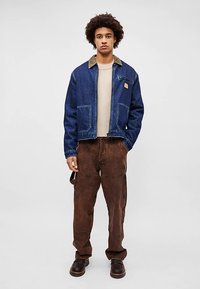 Casaco de ganga azul com gola em veludo cotelê castanha e bolsos frontais, usado sobre uma camisa de malha bege e calças castanhas com acabamento texturizado.