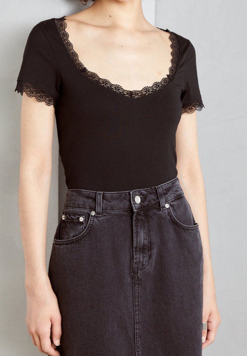 Body nero con pizzo lungo la scollatura e le maniche, abbinato a una gonna in denim scuro a vita alta con tasche frontali e bottoni in metallo.