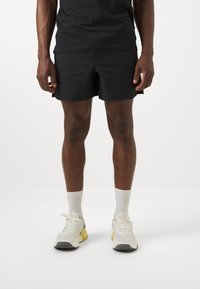 Pantalones cortos negros con un diseño estampado y un aspecto texturizado, combinados con calcetines blancos y zapatillas deportivas de color claro. Camiseta negra lisa utilizada en la parte superior.