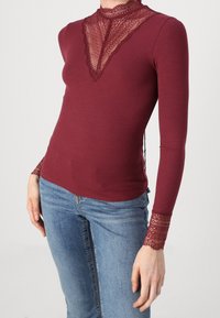 Burgunderfärgad ribbad långärmad topp med spetsdetaljer vid halsringningen och ärmsluten; figurnära silhuett med sidodetalj för extra textur.