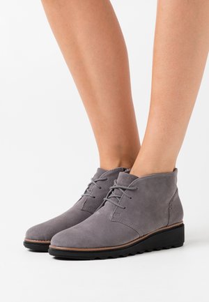 Botines de ante gris con punta redondeada, cordones en la parte delantera y suela de goma texturizada. Presentan un tacón bajo para mejorar la comodidad y el soporte.