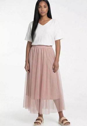 Kobieta o długich czarnych włosach, ubrana w biały krótki top z dekoltem w serek, różową warstwową maxi spódnicę z tiulu i beżowe sandały, stojąca na białym tle.