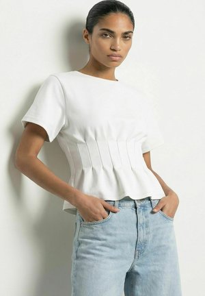 Kvinde iført en hvid tætsiddende kortærmet top med plisseringer og lyseblå højtaljede jeans, stående med hænderne i lommerne.