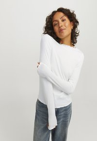 White ribbed long-sleeve top featuring a scalloped neckline and elongated sleeves, paired with blue high-waisted jeans.