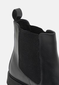 Bottine noire avec un extérieur en cuir lisse et un panneau élastique tissé texturé. Comprend une languette pour un enfilage facile.