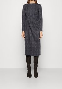 Robe à carreaux gris avec des manches longues, un décolleté carré et un détail twist sur le côté. Associée à des bottes en daim marron arrivant au genou.