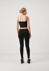 Žena s kučeravými vlasmi zviazanými do drdolu, oblečená v čiernom krátkom topi s tenkými ramienkami, čiernych priliehavých džínsach a čiernych topánkach na podpätku, otočená chrbtom.