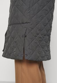 Cappotto trapuntato grigio scuro con spacco all'orlo, dotato di bottoni a pressione. Il tessuto ha una consistenza liscia e leggermente lucida. Combinato con pantaloni marroni.