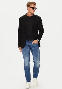 Blazer nero, t-shirt nera, jeans azzurri chiari e sneakers bianche. Gli occhiali da sole e l'orologio al polso completano l'outfit. Aspetto casual ma curato.