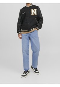 Svart varsityjacka med läderärmar, med en stor vit "N"-logotyp, randig krage och ärmslut, bärs över en beige skjorta och blå jeans.