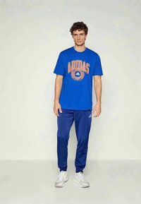 Blå bomulls t-skjorte med oransje "ADIDAS"-logo, kombinert med matchende blå bukser med hvite detaljer og teksturert stoff. Hvite joggesko.