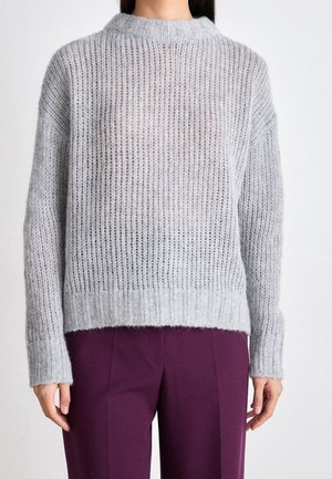 Femme portant un pull en maille côtelée gris clair et un pantalon violet foncé, debout avec les bras détendus le long du corps sur un fond blanc.
