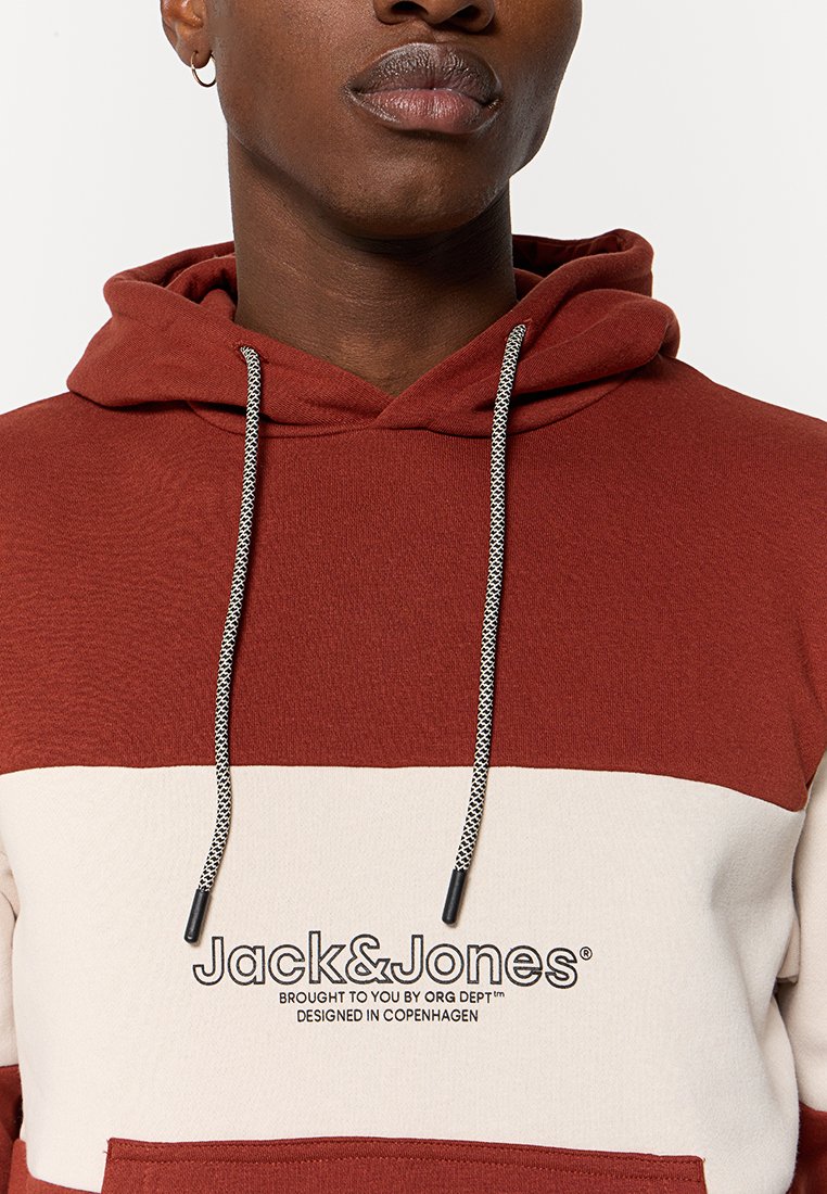 Sweatshirt à capuche rouge et crème avec des cordons de serrage, comportant un logo sur la poitrine. Texture douce, design bicolore et détails en matériel contrastant.