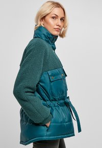 Veste bleu-vert comprenant un haut en polaire doux et un bas lisse et rembourré. Comprend une taille ceinturée, des poches avant et des fermetures à pressions.