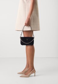 Sort tekstureret clutch taske med en kædestrop og logoaccent. Tasken har en lynlås lukning og holdes i en hånd. Lys rutig kjole.