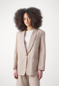 Blazer in lino beige con revers a punta, un bottone dorato e tasca sul petto. La vestibilità ampia e il tessuto testurizzato accentuano il suo design rilassato.