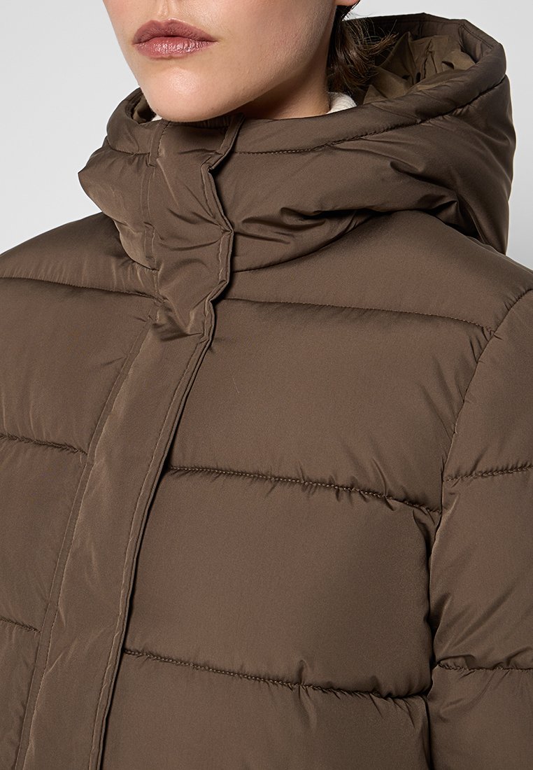 Donna che indossa una giacca invernale imbottita marrone con colletto alto e cappuccio, labbra visibili, su sfondo semplice.