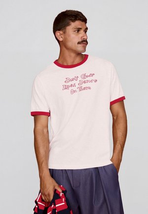 Sötét hajú, bajszos férfi, fehér pólóban, piros szegéllyel, rajta a felirat: „Don't Cross Lines, Dance On Them”, kezében összehajtott piros és tengerészkék csíkos szövetet tart.