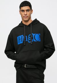 Uomo che indossa un hoodie nero con testo blu "Hugo", mani in tasca, pantaloni neri e una cintura nera, in piedi su uno sfondo bianco.