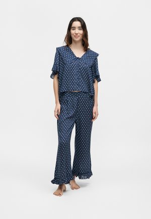 Νεαρή γυναίκα στέκεται ξυπόλητη, φορώντας σετ πιτζάμα μπλε ναυτικού με πουά, με φαρδιά γιακά με βολάν, κοντά μανίκια και ζώνες με βολάν στα παντελόνια.