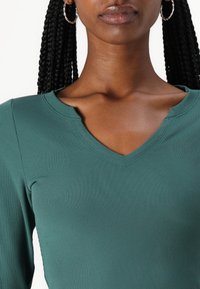 Ribbestrikket topp med v-hals i en mørkegrønn farge med en tettsittende design. Har teksturert stoff og en liten variasjon i halslinjen.