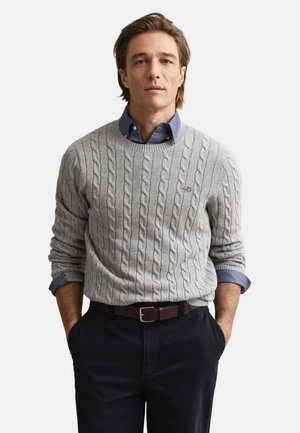 Gray cable-knit sweater with a round neckline, worn over a blue button-up shirt. Dark pants and a brown belt complete the outfit.