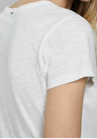 Bílá, krátkosrstá, strukturovaná košile s kulatým výstřihem a viditelným švem. Obsahuje malou značkovou nálepku na zadní straně.