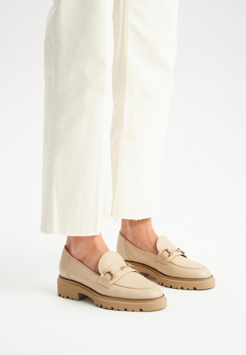 Mocassini beige con tomaia in pelle testurizzata, suola piatta in gomma e dettaglio metallico decorativo. Indossati con pantaloni bianchi a gamba larga e cropped.