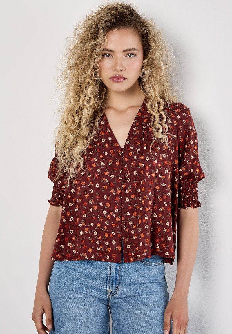 Blusa floreale bordeaux con maniche corte elastiche, scollatura a V e chiusura con bottoni. Il motivo presenta piccoli fiori rossi e crema su un tessuto testurizzato.