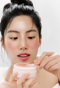 Produit de soin de la peau dans un pot rond rose tenu par une personne. La personne a un teint lisse, des joues roses et applique un produit avec une spatule.