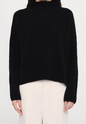 Personne portant un pull noir à col roulé manches longues et un pantalon blanc taille haute, debout, les bras détendus le long du corps.