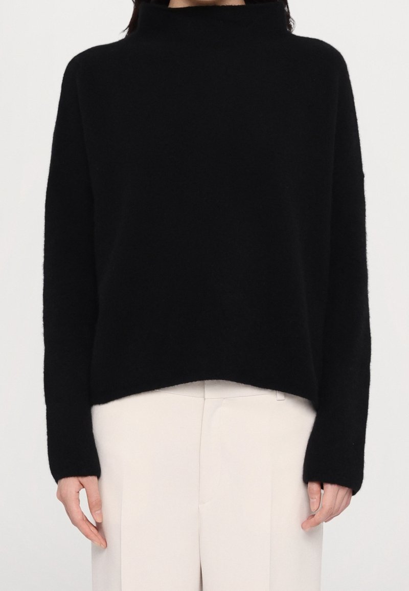 Personne portant un pull noir à col roulé manches longues et un pantalon blanc taille haute, debout, les bras détendus le long du corps.