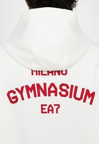 Biely mikina s kapucňou s červeným vyšívaným textom "MILANO GYMNASIUM" a logom EA7 na chrbte, vyrobená z mäkkej, textúrovanej tkaniny.
