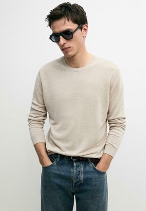 Mężczyzna w czarnych okularach przeciwsłonecznych, beżowym swetrze z długim rękawem i niebieskich dżinsach, z rękami w kieszeniach, na jednolitym białym tle.